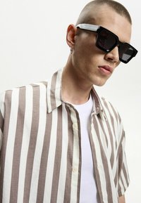 Striped short-sleeve shirt in beige and white with a collar; worn over a plain white T-shirt. Black sunglasses with thick frames.