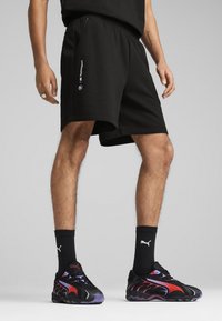 Sorte shorts med elastisk talje og sidelommer, med et BMW Motorsport-logo på venstre side. Båret med sorte og røde sneakers.