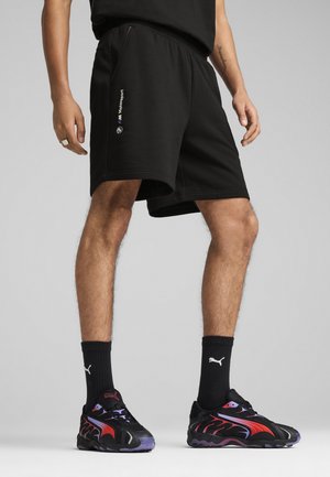 Schwarze Shorts mit elastischem Bund und seitlichen Taschen, mit einem BMW Motorsport-Logo auf der linken Seite. Getragen mit schwarz-roten Sneakers.