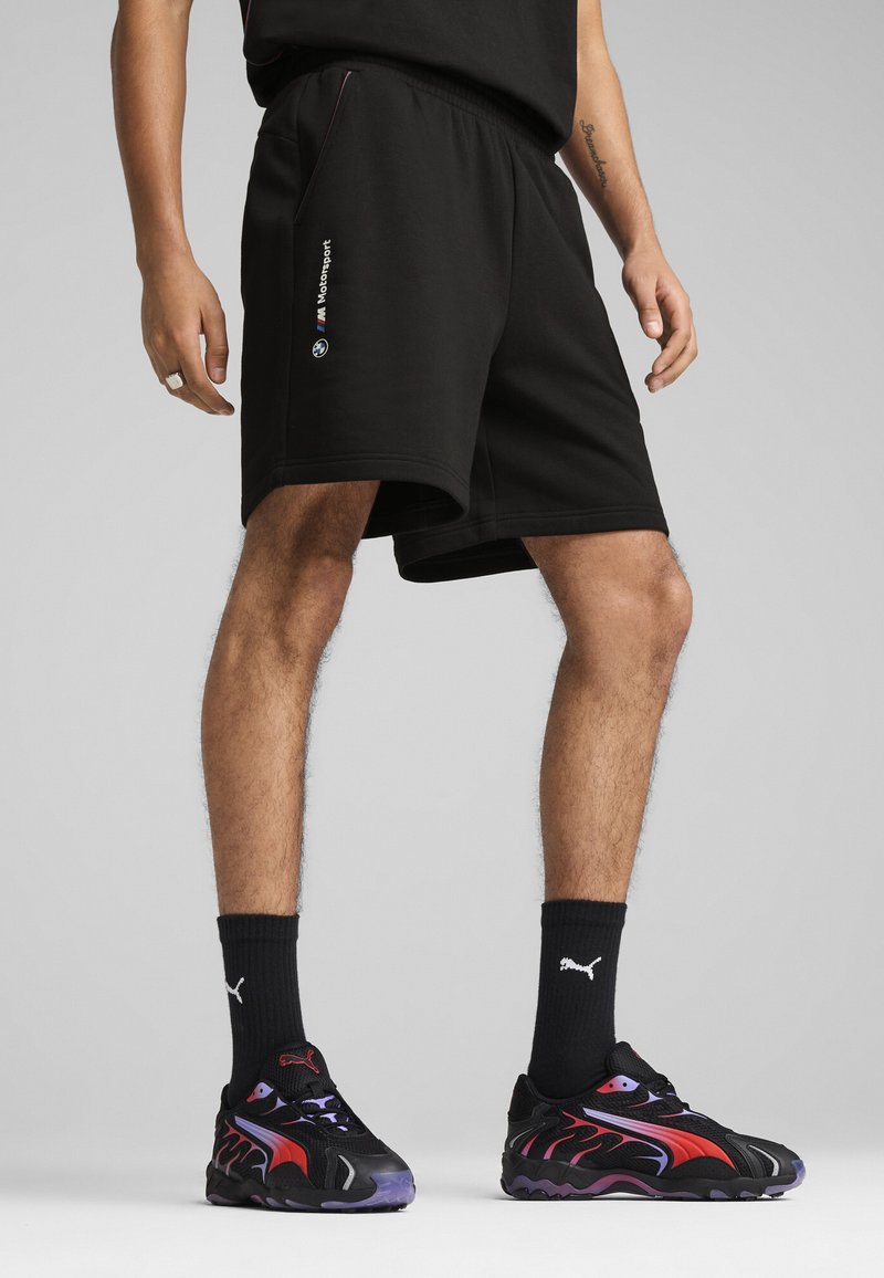 Sorte shorts med elastisk talje og sidelommer, med et BMW Motorsport-logo på venstre side. Båret med sorte og røde sneakers.