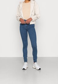 Valkoinen corduroy-tyylinen vetoketjullinen takki, beige ribattu paita, siniset skinny-farkut, harmaat ja valkoiset lenkkarit. Vapaa-ajan asu, jossa muotoillu rakenne ja tiukka design.