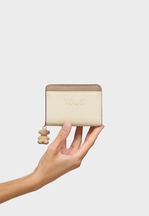 Mano che tiene un portafoglio TOUS beige e marrone con un piccolo ciondolo a forma di orsetto come cursore della cerniera, su uno sfondo chiaro e uniforme.