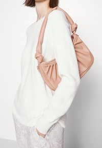 Sac à main en cuir rose doux avec une anse nouée, présentant un design froncé et une fermeture éclair visible, porté sur un pull blanc.
