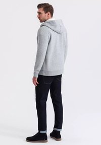 Felpa grigia con cappuccio e cordino, tessuto strutturato, tasca a marsupio, abbinata a jeans scuri e stivali in suede neri, in piedi su uno sfondo bianco.