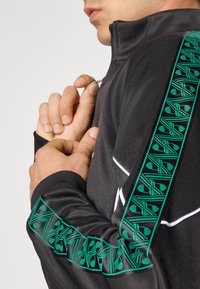 Gros plan sur une personne qui ferme une veste noire avec des rayures vertes à motif et des bordures blanches sur la manche et l'épaule.