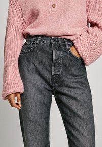 Suéter de punto color rosa claro con un patrón texturizado y un solo botón, combinado con unos vaqueros de denim gris oscuro de tiro alto que cuentan con bolsillos frontales.