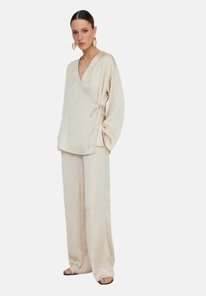 Kvinde iført løs beige slå-om-bluse og bukser med vidde ben, med store guldløv-øreringe og sandaler, stående mod en hvid baggrund.