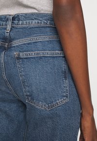 Siniset farkut korkealla vyötäröllä, joissa on näkyvät takataskut ja kontrastitiksaukset. Kangas on klassinen ja kestävä.