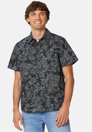 Mann trägt schwarzes kurzärmeliges Hemd mit grauem Blumenmuster und hellblauen Jeans, lächelt und steht vor einem einfarbigen Hintergrund.