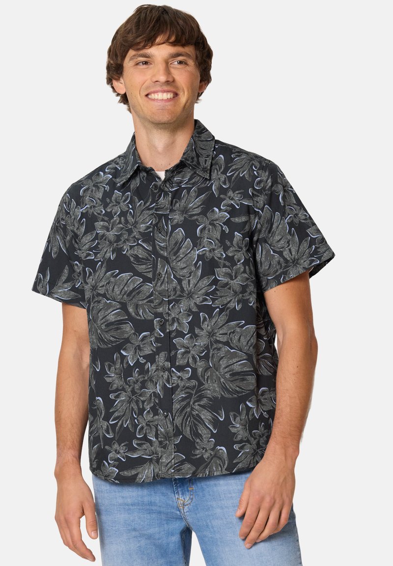 Homme portant une chemise noire à manches courtes avec un imprimé floral gris et un jean bleu clair, souriant et debout devant un fond uni.