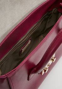 Bordeaux läderhandväska med en slät yta, inre fack och detaljer i guldfärg. Öppen topp som avslöjar en beige foder.