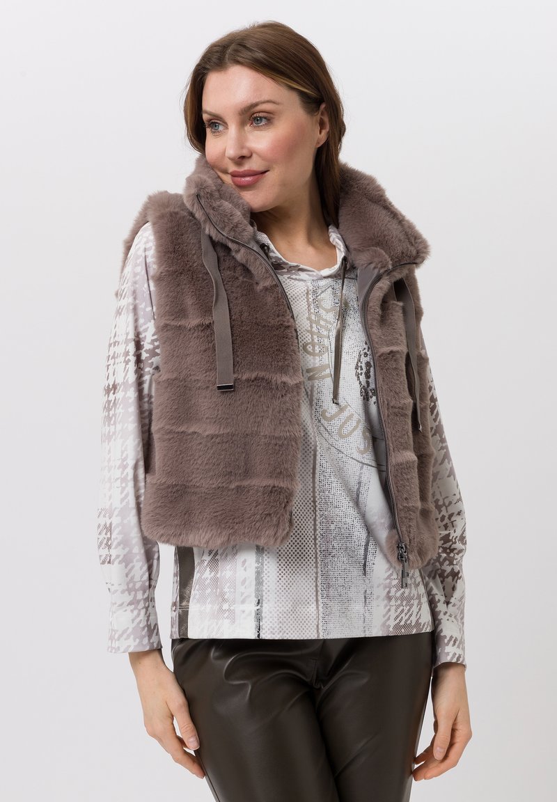 Brown faux fur vest with a hood, featuring a zip front and drawstring. Worn over a patterned long-sleeve top and dark pants.