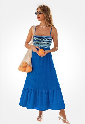 Blauwe maxi-jurk met een patroon op het lijfje, een gekreukte textuur en een gerimpelde zoom. Draagt heldere sandalen en houdt sinaasappels vast in een netzak.