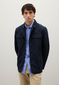 Junger Mann in einer marineblauen Jacke über einem blau-weiß gestreiften Hemd mit Knöpfen und beigen Hosen, der vor einem schlichten hellen Hintergrund steht.