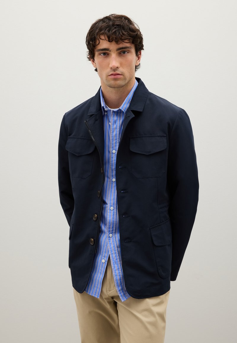 Junger Mann in einer marineblauen Jacke über einem blau-weiß gestreiften Hemd mit Knöpfen und beigen Hosen, der vor einem schlichten hellen Hintergrund steht.