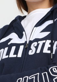Marinblå zip-up hoodie med vitt logo och text. Mjuk tissu, känguruficka, dragsko på huven och ribbad manschett. Klassisk passform.