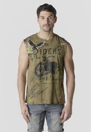 Hombre con camiseta sin mangas verde oliva con gráficos de motocicleta y águila, combinado con shorts cargo de mezclilla clara, de pie contra un fondo gris.