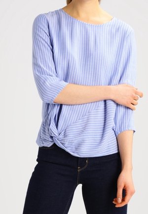 Femme portant un haut rayé bleu clair et blanc avec des manches trois-quarts noué en nœud à la taille et un jean bleu foncé.