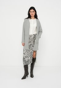 Γκρι μακρύ ζακέτα πάνω από λευκό πουκάμισο με κουμπιά, συνδυασμένη με ασπρόμαυρη φούστα με σχέδιο και σκίσιμο, και μαύρες μπότες μέχρι το γόνατο.