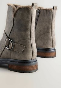 Graue Wildleder-Stiefeletten mit Kunstpelzbesatz, seitlichem Reißverschluss und silberner Schnalle. Robuste Gummisohle mit braunem holzähnlichem Akzent.