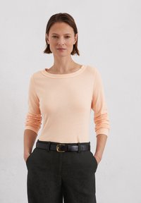 Hellpfirsichfarbener langärmliger Pullover mit weitem Bootsausschnitt, kombiniert mit dunklen Hosen und einem schwarzen Gürtel mit einer goldenen Schnalle.
