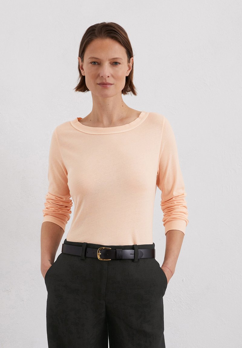 Hellpfirsichfarbener langärmliger Pullover mit weitem Bootsausschnitt, kombiniert mit dunklen Hosen und einem schwarzen Gürtel mit einer goldenen Schnalle.