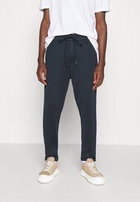 Marineblaue Jogginghosen mit Kordelzugbund, Seitentaschen und schmalem Schnitt. Kombiniert mit beigen Sneakers, die weiße Schnürsenkel und eine Gummisohle haben.