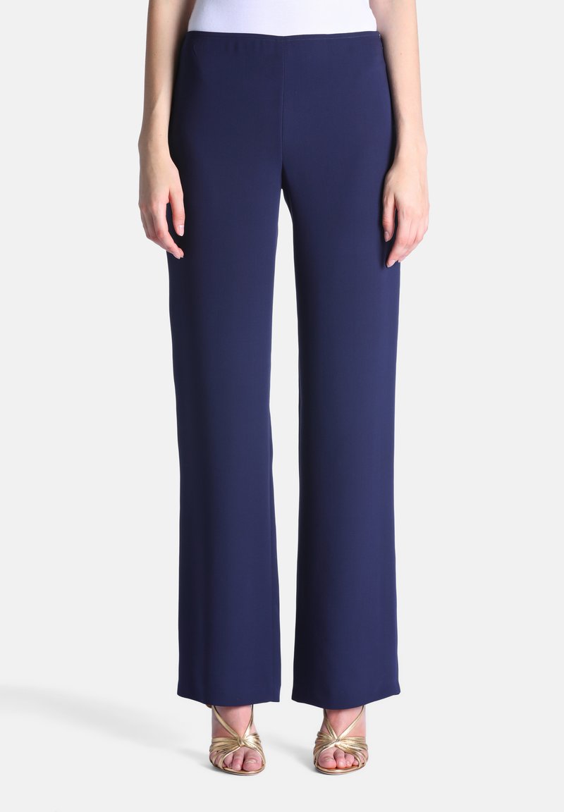 Femme portant un pantalon ample bleu marine associé à des sandales ouvertes à talons hauts dorées, debout devant un fond uni.