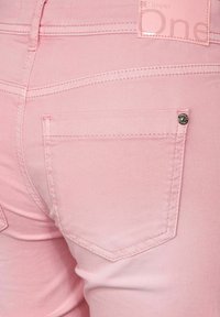 Gros plan de la poche arrière d'un pantalon en jean rose clair avec des coutures visibles et un petit rivet métallique dans le coin supérieur droit de la poche.