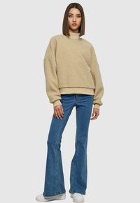 Pull en polaire beige avec épaules tombantes et col montant, associé à un jean en denim flair et des baskets blanches. Design simple et texturé.