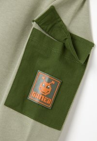 Un bolsillo de algodón verde en una camiseta gris claro presenta un parche naranja con la palabra "GAMER" y un diseño de personaje estilizado. La tela tiene una textura suave.