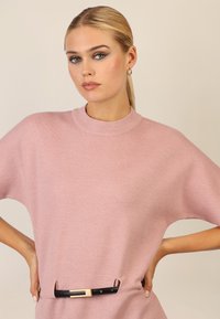 Helle rosa Sweatshirt mit hohem Kragen und kurzen Ärmeln, versehen mit einem schwarzen Gürtel mit einer metallischen Schnalle in der Taille. Strukturierter Strickstoff.