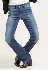 Blå denim jeans med utsvängd ben är i mellan tvätt, har framfickor och synliga sömmar. Kombineras med svarta klackade ankelstövlar.