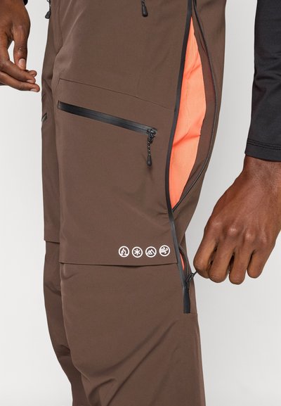 Bruna byxor med dragkedja, svarta detaljer och orange innertyg. Har en dragkedjeförsedd ficka och fem logotypikoner på benet.