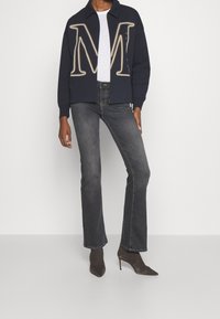 Giacca a maglione blu navy con grande logo "M" beige e t-shirt bianca sotto, abbinata a jeans grigi a zampa d'elefante e stivali neri con tacco alto.