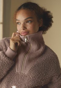 Texturovaný, plyšový růžový pulovr s vysokým límcem a zlatým zdrhovadlem. Vyznačuje se žebrovanými a nadýchanými materiály, zvýrazněný přiléhavým střihem.