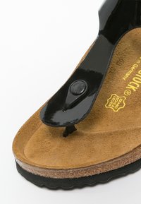 Svart patenterad tongsandal med korkfotbädd, tan mocka textur, gummisula och präglad logotyp på innersulan.