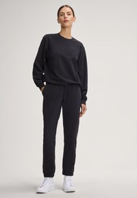 Completo sportivo nero composto da una felpa con collo rotondo, caratterizzata da un logo in rilievo, e pantaloni jogger abbinati con polsini elasticizzati, abbinato a sneakers bianche.