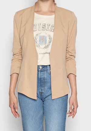 Frau trägt einen beigen offenen Blazer über einem weißen grafischen T-Shirt und blauen hoch taillierten Jeans, stehend vor einfachem Hintergrund.