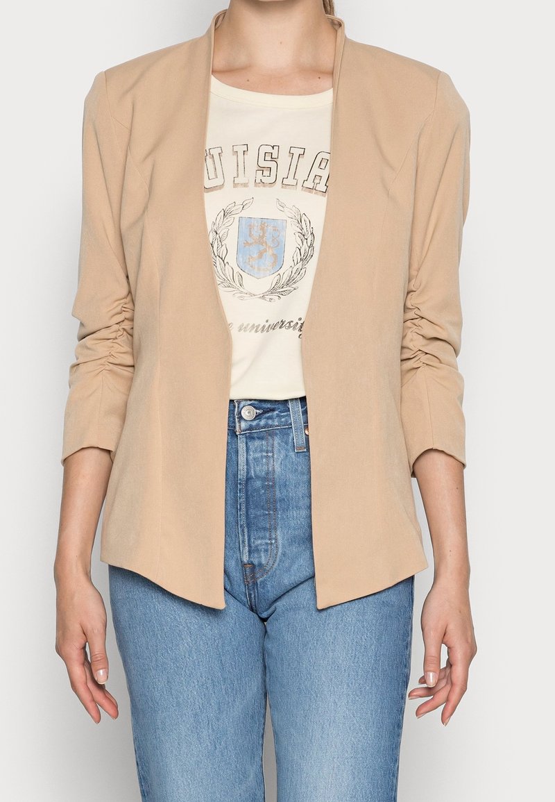 Femme portant un blazer beige ouvert devant sur un T-shirt blanc à motif graphique et un jean bleu taille haute, debout devant un fond uni.