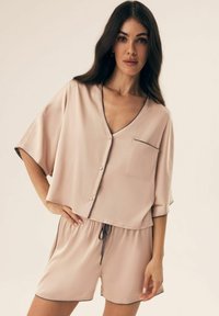 Kobieta z długimi ciemnymi włosami, ubrana w beżową satynową bluzkę z guzikami i pasujące szorty z ciemnym obszyciem, stoi z jedną ręką na biodrze.