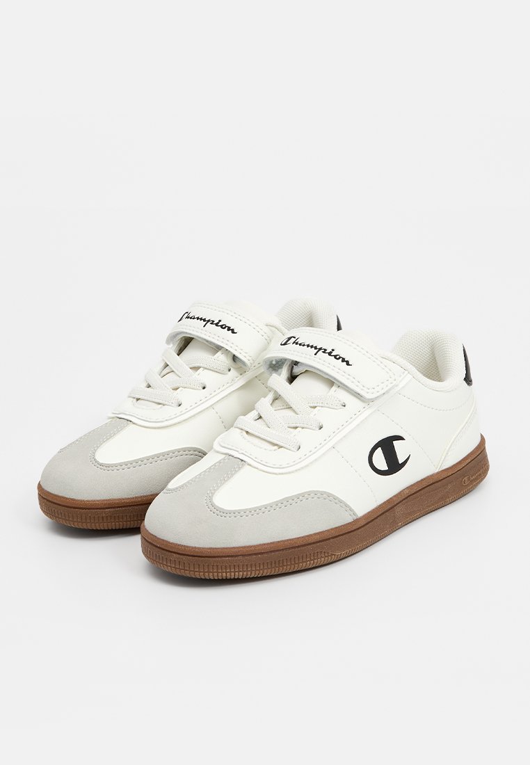Zapatilla blanca con acentos de ante gris, suela de goma, cierre de cordones y tira de velcro. Presenta el logotipo "Champion" en negro en el lateral.