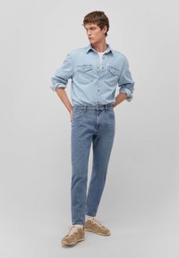 Camicia di jeans azzurro chiaro con design a bottoni, due tasche sul petto, abbinata a jeans azzurro chiaro e sneakers beige. Il modello sta in piedi con le mani sui fianchi.
