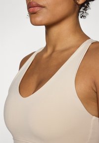 Beige sportsbh med dyp V-halsdesign, laget av et glatt, elastisk stoff. Tynne stropper og minimalistiske sømdetaljer.