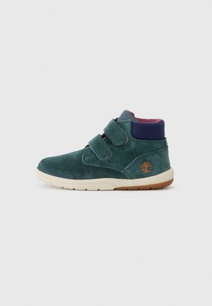 Bottines hautes en suede vert avec deux straps en Velcro, semelle cream, col rembourré et logo brodé sur le talon.