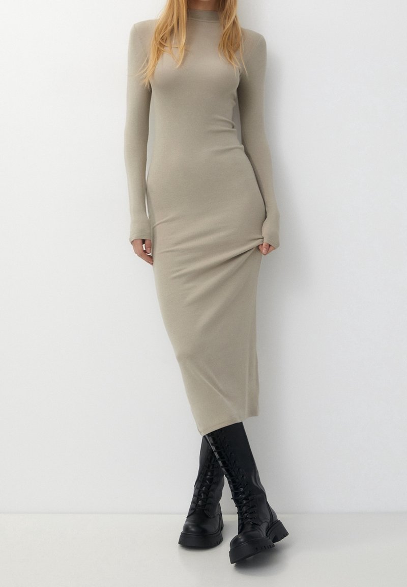 Femme portant une robe midi beige cintrée à manches longues et des bottes noires montantes à lacets sur un fond blanc uni.