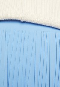 Gonna plissettata azzurra chiaro realizzata in tessuto liscio, sovrapposta a un top lavorato a maglia color crema con texture a coste, che mostra uno stile casual.