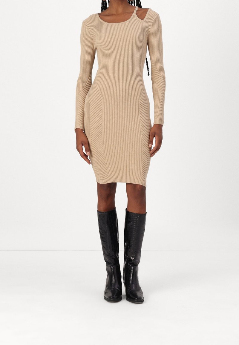 Femme portant une robe en maille côtelée beige à manches longues avec une découpe près de l'épaule gauche, associée à des bottes noires montant jusqu'aux genoux.
