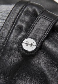 Borsa nera in pelle con bordi cuciti e un bottone metallico circolare che presenta la scritta "PEARLWOOD". La superficie è liscia con una leggera lucentezza.