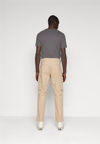 Pantalones cargo beige con múltiples bolsillos laterales, hechos de un material de algodón duradero, combinados con una camiseta gris lisa, vistos desde la parte trasera.